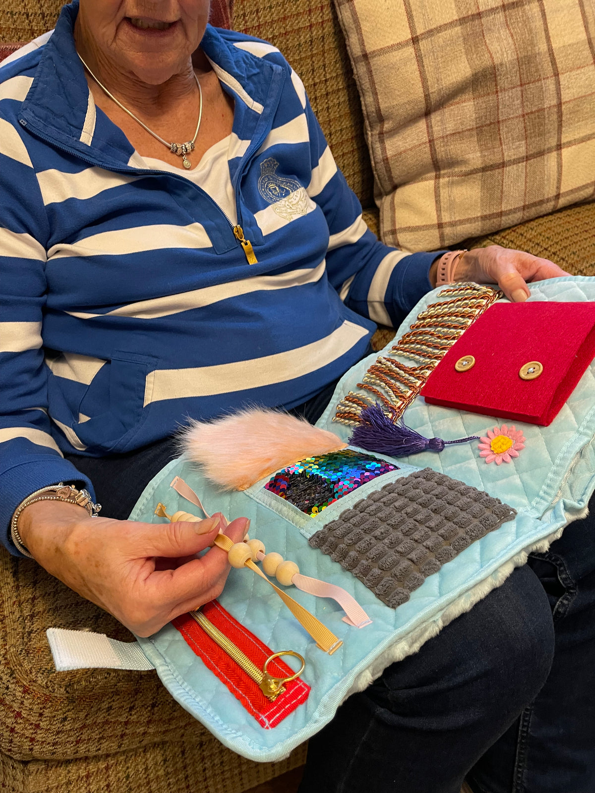 Eldre dame i en sofa fikler med perler og tekstiler på et lyseblått fikleteppe i fanget